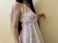 Tisia wearing a white lace and sheer babydoll with her tits partially visible through the fabric, long dark hair, standing against a plain wall.