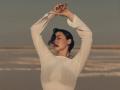 OliviaCluff with arms raised, eyes closed, wearing a sheer white long-sleeve top, short dark hair, soft light on her face, gentle beach background, relaxed, calm expression.
