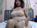 FelyLove, a curvy woman with dark brown hair, sits on a bed wearing a sheer, beige dress that reveals her large tits and nipples, with her legs crossed.