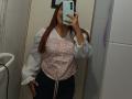 AntoniaBrunni with long brown hair, wearing a white blouse with pink floral pattern and black pants, smiling in a mirror selfie, holding a blue phone, showing her tits.