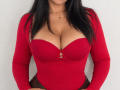 CharlotteDevereaux with large tits in a red, long-sleeve, low-cut top, revealing ample cleavage, black hair, and a small gold necklace against a plain white background.