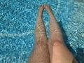 Barbarica's legs in a blue-tiled pool, water rippling over her skin, toes pointed upwards, reflecting sunlight. Her thighs are slightly apart, showing a bit of her ass.