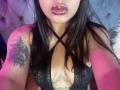 ChiaraBella with glossy pink lips, black lace top, crisscross straps, showing cleavage, tattoo on right arm, long black hair, pink and purple light.