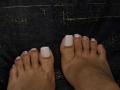 PaulethJoens' feet with white, square-tipped nails, slight gold glitter on the tips, resting on a dark textured surface.