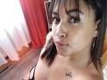 MirandaDreams with brown hair and bangs, pouty lips, light brown skin, small tattoo on chest, black strap top, gold earrings, standing in a room with red and white curtains.