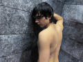 RubyMote with wet black hair, bare back, and small ass, leaning against dark gray textured tiles, looking over shoulder. Skin is pale, water droplets visible.