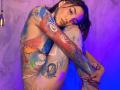 LunaixMoon is naked with colorful paint on her tits, ass, and arms, hugging her knees. She has tattoos, dark hair, and a purple-blue light background. Photographic style.