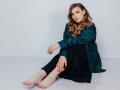 Niferttiti with wavy brown hair, green satin jacket, black pants, bare feet, red nails, sitting on white floor, arms folded, looking at camera, soft expression, full lips.