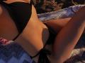 Olieri's sunlit body in a black bikini, showing her small, perky tits and slim, toned ass, lying on a textured blanket with her legs slightly apart.