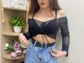 Rioria with big boobs, wearing a sheer black top, exposing her tits, and blue jeans with a black belt, pouty lips, and long brown hair, standing in a room.