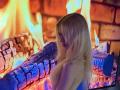 Dierdre Lorenzano, avec ses longs cheveux blonds, jetant un coup d'œil par-dessus son épaule, se tient devant un feu aux flammes bleues et oranges, montrant son dos nu et un petit tatouage..