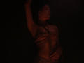 TriciaBarnes with dark red rope binding her tits and chest, arm raised, in dim, moody lighting. Her hand rests on her crotch. Her face is turned to the side.