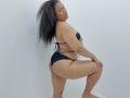 KloheEvans with curly black hair, wearing a black bikini, kneels on one leg, showing her ass, holding her tits, looking back with a pouty face.