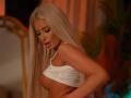 LaraBrynn with long platinum blonde hair, wearing a white tank top, exposing her tits, looking down with her mouth slightly open, in a warmly lit room.