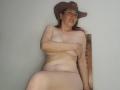 MadisonDurand is nude, sitting with a brown cowboy hat, covering her tits with one arm, smiling, and showing a bit of her ass, against a plain white wall.