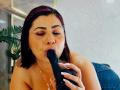 AngeleRouxo, with black hair and red headband, is sucking on a large black dildo, eyes closed, wearing denim and a ring, in a provocative, intimate moment.