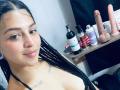 LonxEva with braided black hair, light brown skin, and small tattoos, smiling at the camera, showing her bare shoulders and black strap, with a shelf of dildos in the background.