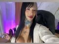 AnneKisss with black hair, white shirt, tattoos on neck and chest, pink eyeshadow, red lipstick, holding a phone, purple and pink lighting, black chair in background.