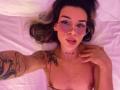 LenaBranch with pink-tinted glasses, dark blonde hair, tattoos on arm and chest, freckles, looking at camera, hand on chest, lying on white bed, small pink bra.
