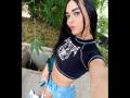 SexyHannaD with long black hair, wearing a black crop top with white design, blue high-waisted shorts, holding a white bag, has tattoos, hoop earrings, and pink lipstick.