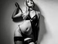 MalloryHearth with blonde hair, wearing black lace lingerie, holding a strap, showing her tits, ass, and a sexy smile, standing against a wall in a black-and-white photo.