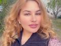 AvaQueen has curly blonde hair, green eyes, and pink lips. She's wearing a black zip-up vest with pink floral sleeves. She's sporting small, gold earrings.