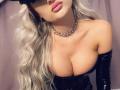 MademoiselleBella with platinum blonde wavy hair, black mask, pink lips, shiny black gloves, and a silver choker, showcasing her large, glossy tits in a tight, black latex top.