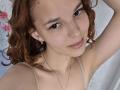 ChayaParrett with curly red hair, small tits, fair skin, and a slight smile, wearing a beige top, has a gold necklace, and small earrings, looking at the camera.