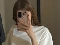 Fotografía de Calista Swiatek, con cabello castaño largo, camiseta blanca, sosteniendo un teléfono con una funda floral que le cubre parcialmente el rostro, de color oscuro. Uñas rojas, tomando una foto con el móvil, tensión sexual sutil, ambiente informal..