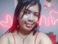 MariahSquirting69, con piel brillante, lápiz labial rojo y cabello negro, vestida con una camiseta roja de tirantes finos, collar con una piedra rosa, luces de corazón neón en el fondo, sonriendo a la cámara..