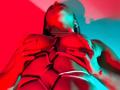 MelodyRoss with red and blue lighting, holding her large tits in a strappy black harness, fingers gripping her chest, shadowed face, long dark hair, ring on left hand.