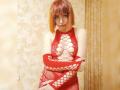 LaylaHottyX with orange bob, red lace-up dress, and matching arm sleeves, showing off her tits through the mesh, standing with arms crossed, smiling subtly.