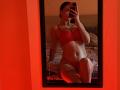 Olieri in red lingerie, sitting in front of a mirror, showing tits and pussy, holding a phone, red room, slight shadow on bed, reflection, dark hair, red light.