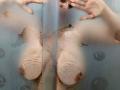 Reflejo de los senos grandes, pálidos y desnudos de AnyelinaMilf, y de sus pezones pequeños y marrones, a través de un vidrio sucio y rayado. Manos con uñas cortas tocando el vidrio..