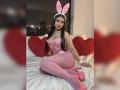 MiilaWx, a sexy woman with long black hair, wears a pink corset, pink stockings, and bunny ears, sitting on a bed with red hearts, her legs spread, showing a flirty vibe.