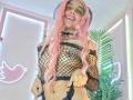 GabySmiith with pink hair, wearing a black fishnet top, black choker, and black thong, smiling at the camera, showing pierced navel, and pointing forward.