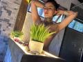 Topless LiaMiss with arms raised, tattooed chest, and short dark hair, standing in front of a window with potted green plants on a lit stone ledge.