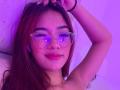 SashaHans with red hair, wearing clear glasses with blue reflections, raised arm, purple light, slightly smirking, visible freckles on her face, pink lipstick, black wristband.