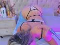 SashaPolet, blonde, in black strappy lingerie, on bed, back to camera, butt up, hand on bed, purple and blue lighting, bed with tufted beige headboard.