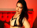 NahomyAllen with long black hair, wearing black lace bra, showing her tits, looking at camera, red tufted headboard in background, red wall, serious expression, warm lighting.