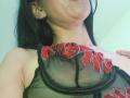 ZarayFeet with black hair, pouty lips, wearing a sheer black bra with red floral embroidery, exposing her titties, her hand on her chest. Light green background.