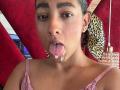 NatalyBlonder with wet, pink tongue hanging out, sticky white fluid on her chin, brown eyes, dark hair in a ponytail, wearing a pink lace top, earrings.
