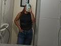 AntoniaBrunni with red hair, wearing a black sheer top and blue jeans, standing in a bathroom, taking a mirror selfie with her phone, showing her curvy ass.