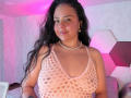 ArielleWilde with long black hair, wearing a pink, see-through, knit top that reveals her big, round tits. She has a pearl necklace and a butterfly earring.