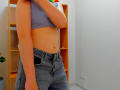 Steisii with a flat stomach, wearing a blue crop top and high-waisted denim jeans, has a visible navel and a slightly raised arm, showing a small tattoo on her side.