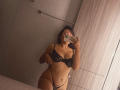 AmeliRoux taking a mirror selfie in a bathroom, wearing black lace lingerie with her tits and pussy visible, holding a phone, in a dimly lit room.