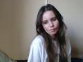 NellaKoczur with long brown hair, white shirt, and pouty expression, slightly blurry, showing her small, subtle tits, and a slight, cute ass, in a casual, indoor setting.