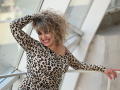 CandyLadyZ with curly blonde hair, wearing a leopard print dress, smiling, hand in hair, standing by a window, showing her tits, and red-painted nails.