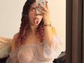 ValeryScot with curly red-pink hair, sticking out tongue, taking mirror selfie, wearing sheer off-shoulder top, showing big boobs, red nails, gold necklace, and bracelets.