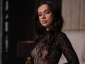 IsadoraReyes with dark wavy hair, wearing a black lace top, showing her tits through the sheer fabric, looking down with a pouty expression.
